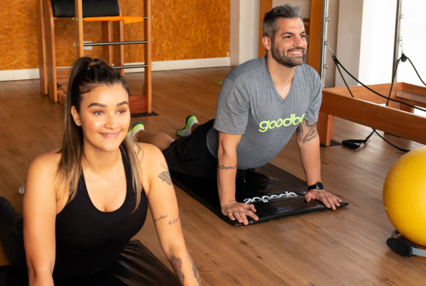 Dois adultos praticando exercícios em uma aula de pilates em um estúdio da Goodbe. A mulher à esquerda está sorrindo enquanto faz uma postura de alongamento, e o homem à direita também sorri, realizando o mesmo exercício em uma posição de prancha. Ambos usam roupas de treino e parecem relaxados e felizes, aproveitando a prática em um ambiente bem equipado, com aparelhos de pilates ao fundo.