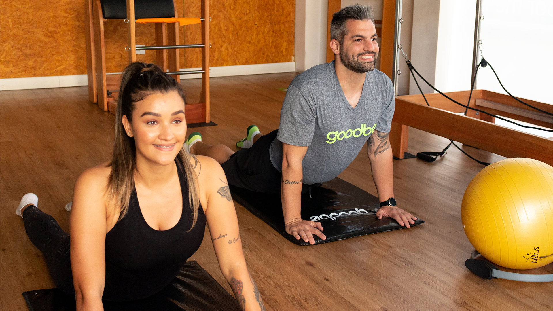 Dois adultos praticando exercícios em uma aula de pilates em um estúdio da Goodbe. A mulher à esquerda está sorrindo enquanto faz uma postura de alongamento, e o homem à direita também sorri, realizando o mesmo exercício em uma posição de prancha. Ambos usam roupas de treino e parecem relaxados e felizes, aproveitando a prática em um ambiente bem equipado, com aparelhos de pilates ao fundo.