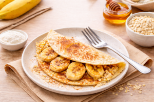 Crepioca de banana em um prato, recheada com rodelas de banana e polvilhada com canela, acompanhada de aveia e mel ao fundo.