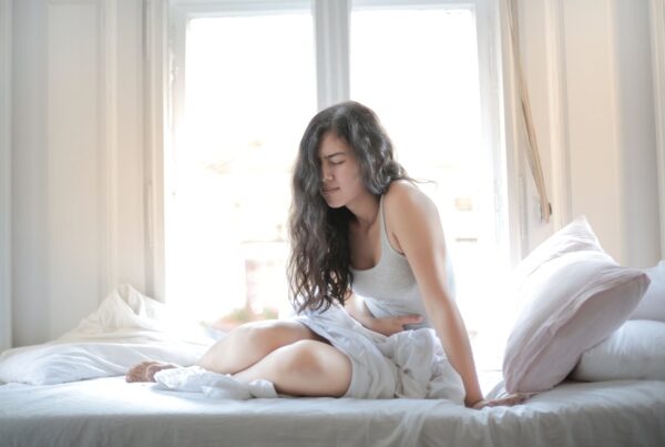 Mulher sentada na cama com expressão de dor abdominal, segurando a barriga em um quarto iluminado