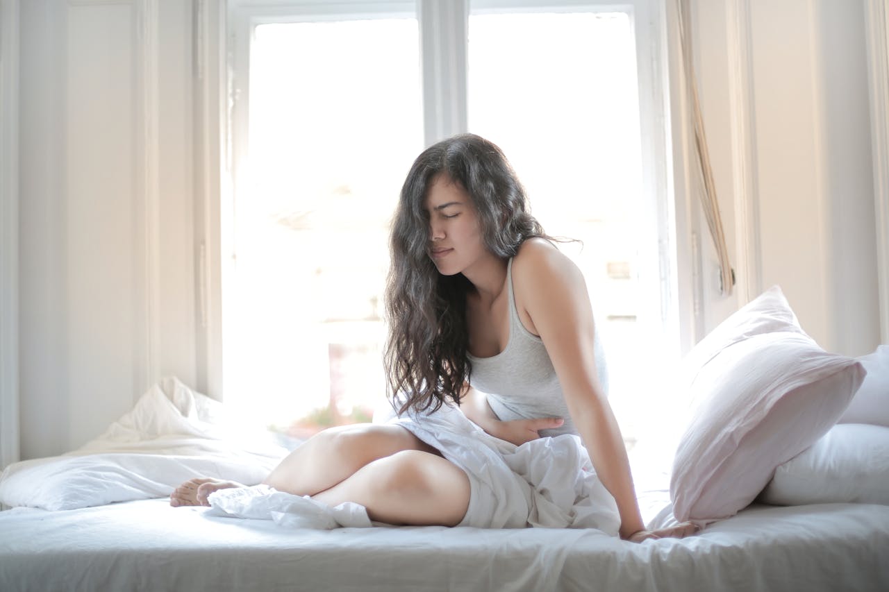 Mulher sentada na cama com expressão de dor abdominal, segurando a barriga em um quarto iluminado